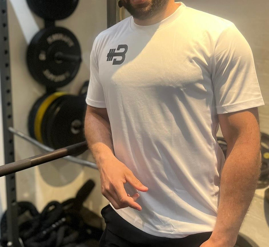 A person wearing a white short sleeve activewear T-shirt with a logo on the chest, in a gym setting with exercise equipment in the background.